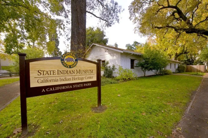 Wooden sign for State Indian Museum with California State Parks logo with large tree and small adobe building in the background