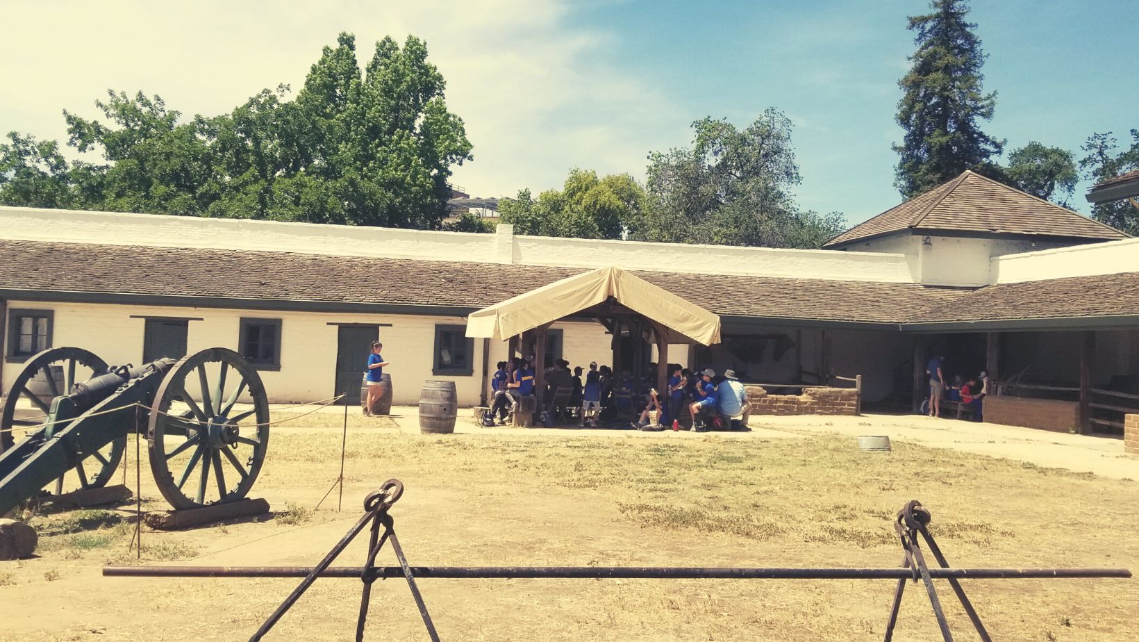 students gather under a wooden ramada inside Sutter's Fort. a green cannon on the left-hand side and firepit in the foreground