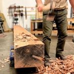 Wooden beam rests on ground with pile of shavings underneath. The legs and arm of a person in California State Parks uniform holds a tool. 
