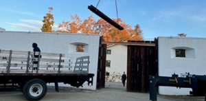 A crane hoists a wooden beam into the sky above the wooden gate at Sutter's Fort. A truck is in the foreground and a figure walks behind the gate. 