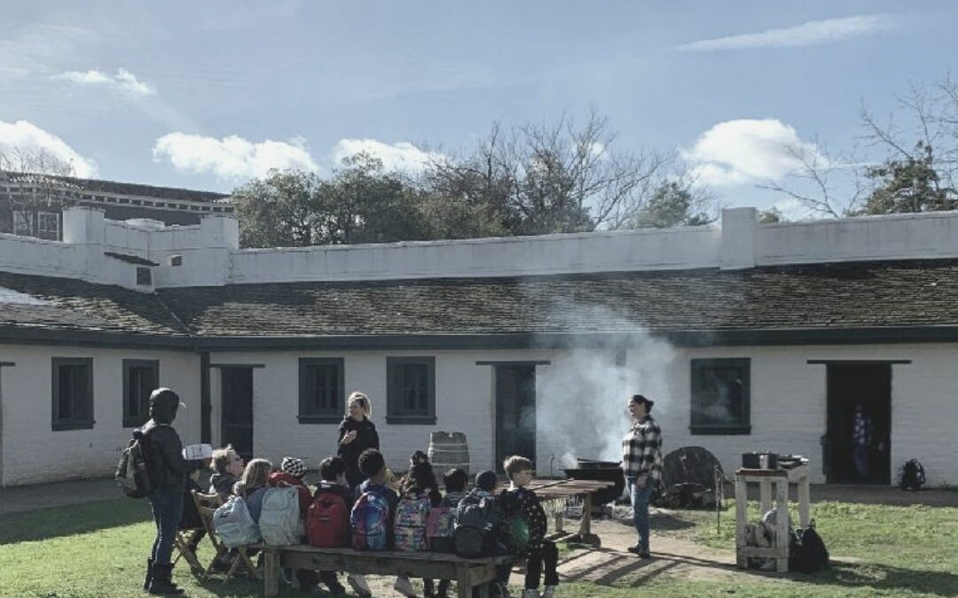 students sit with backs to camera on a bench in the middle of Sutter's Fort. A large pot with steam rising, with an adult standing.