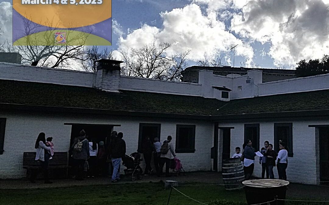 Under a cloudy sky, people explore the historic structures of Sutter's Fort. White adobe brick building with wooden shake roof, patchy grass and dirt ground. a large pot stands on the right-hand side.