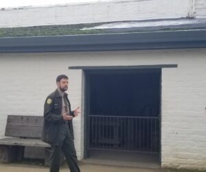 State Park Staff Member talks in front of a room at Sutter's Fort. The roof is covered with tarps and green moss is growing around the shingles.