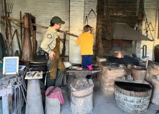 A student works alongside blacksmith inside the blacksmith shop at Sutter's Fort