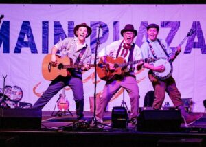 The three musicians from the Rock Bottom Boys on stage holding their string instruments. They appear to be singing and dancing. 