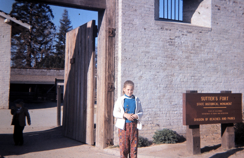Girl stands in front of large wooden gates. Sign for Sutter's Fort State Historical Monument to her left
