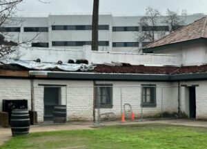 Chain link fence blocks access to white brick building. Wood shake roof is partially exposed, tarps lie on top of roof.