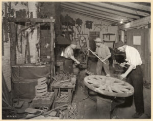 4 men wearing hats and clothing from the early 1900s work in a blacksmith shop. Tools and equipment line the shelves and walls. 