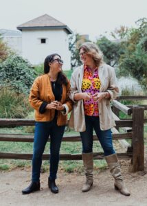 2 women stand in front of wooden fence with sutter's fort bastion in background. Women are looking at each other and laughing. 